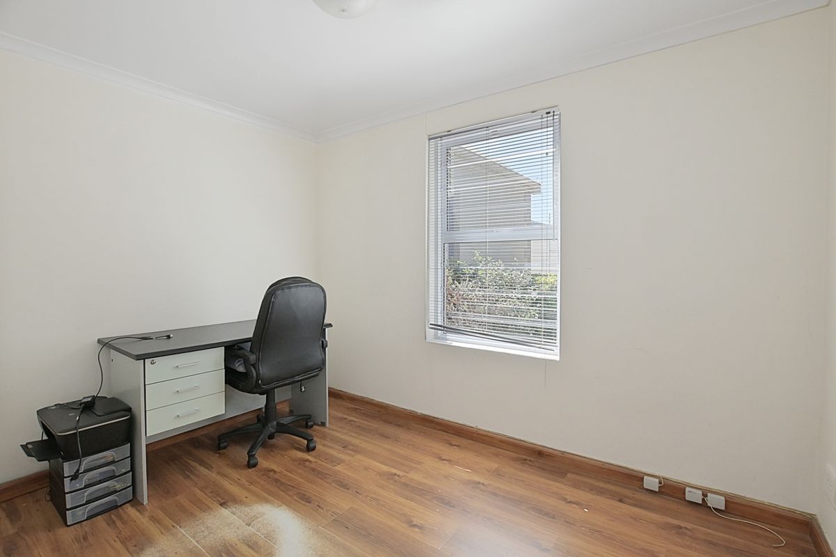 A functional bedroom or home office space featuring laminate wood flooring, a single window with fitted blinds, and neutral wall finishes.