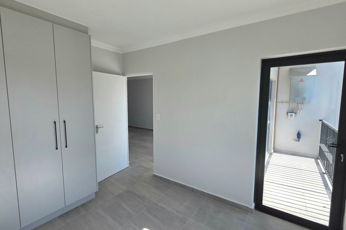A clean, modern bedroom featuring built-in cupboards, neutral tiled flooring, and direct access to a small balcony area.