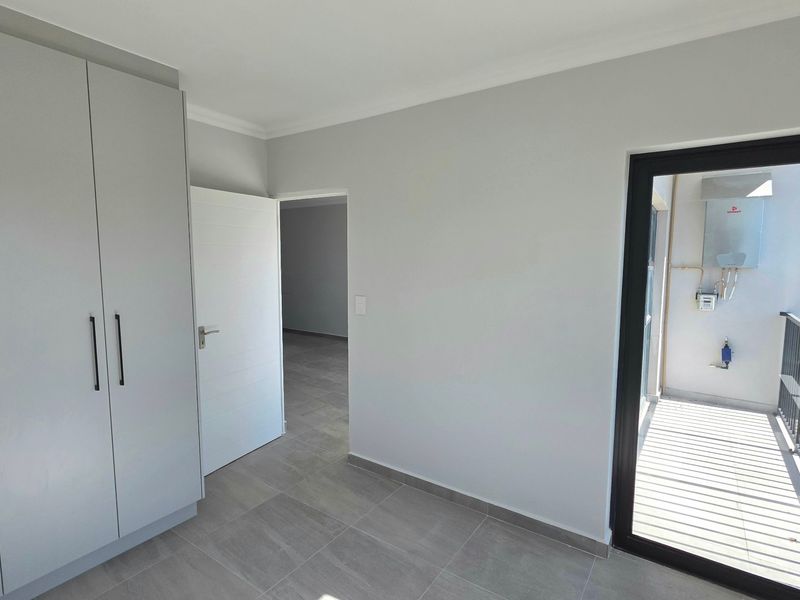 A clean, modern bedroom featuring built-in cupboards, neutral tiled flooring, and direct access to a small balcony area.