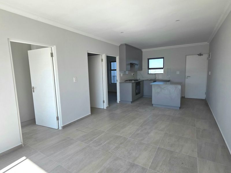 An open-plan living and kitchen area featuring modern grey cabinetry, built-in appliances, a central island, and neutral tiled flooring throughout.