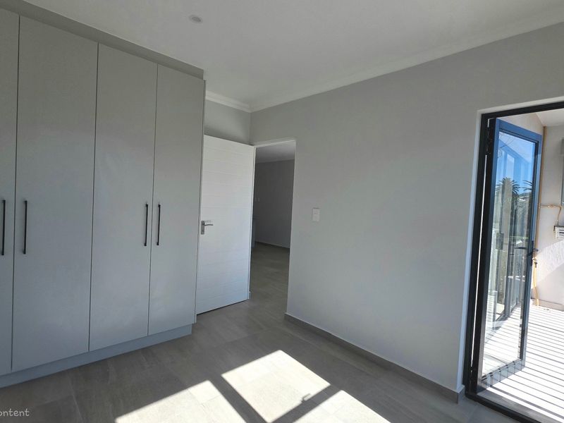 A bright, empty bedroom featuring modern built-in cupboards, neutral tiled flooring, and direct access to an outdoor balcony area.