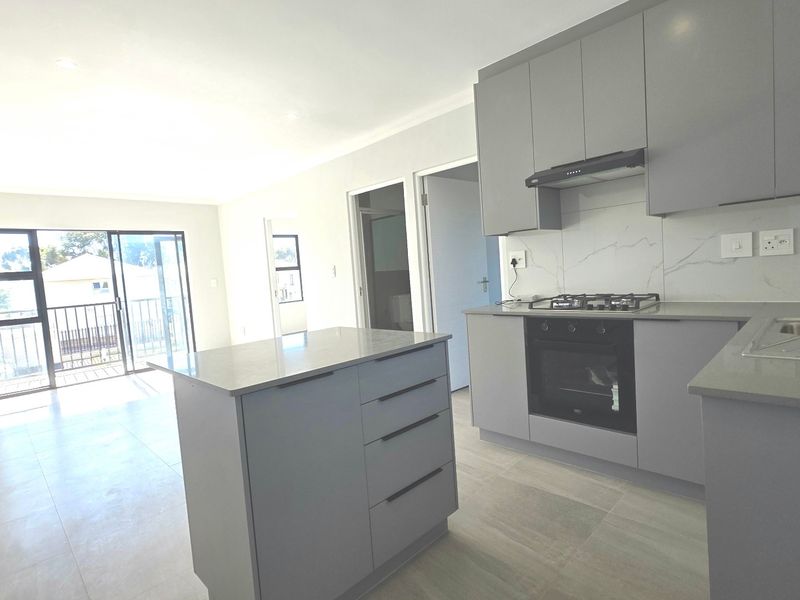 Modern open-plan kitchen and living area featuring sleek grey cabinetry, stone countertops, built-in appliances, and sliding doors leading to a balcony.