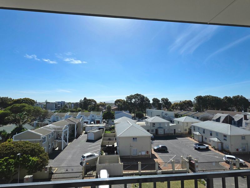 Elevated view overlooking a residential apartment complex with paved parking areas and surrounding greenery under a clear blue sky.