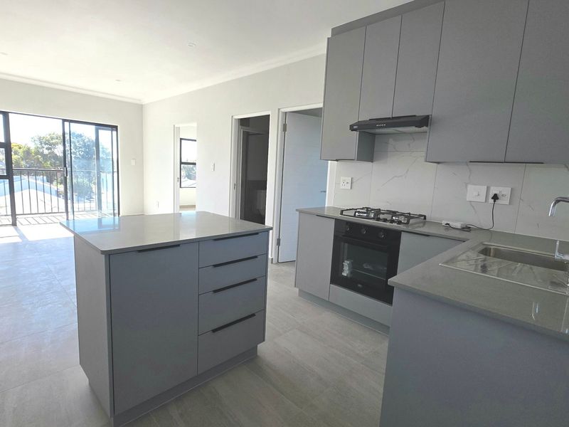 Modern open-plan kitchen and living area featuring sleek grey cabinetry, stone countertops, built-in appliances, and sliding doors leading to a private balcony.