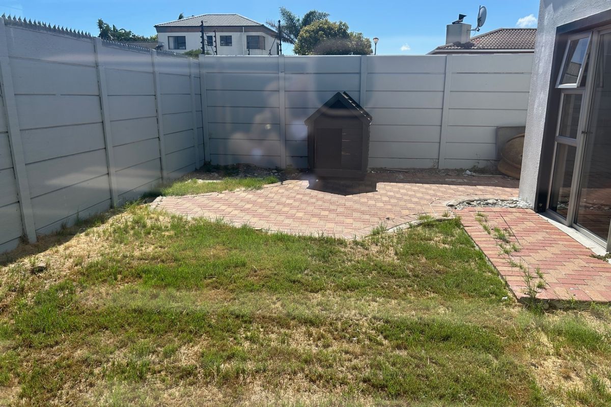 A small rear garden area featuring a paved patio section, perimeter boundary wall, and lawn space, viewed through a window.