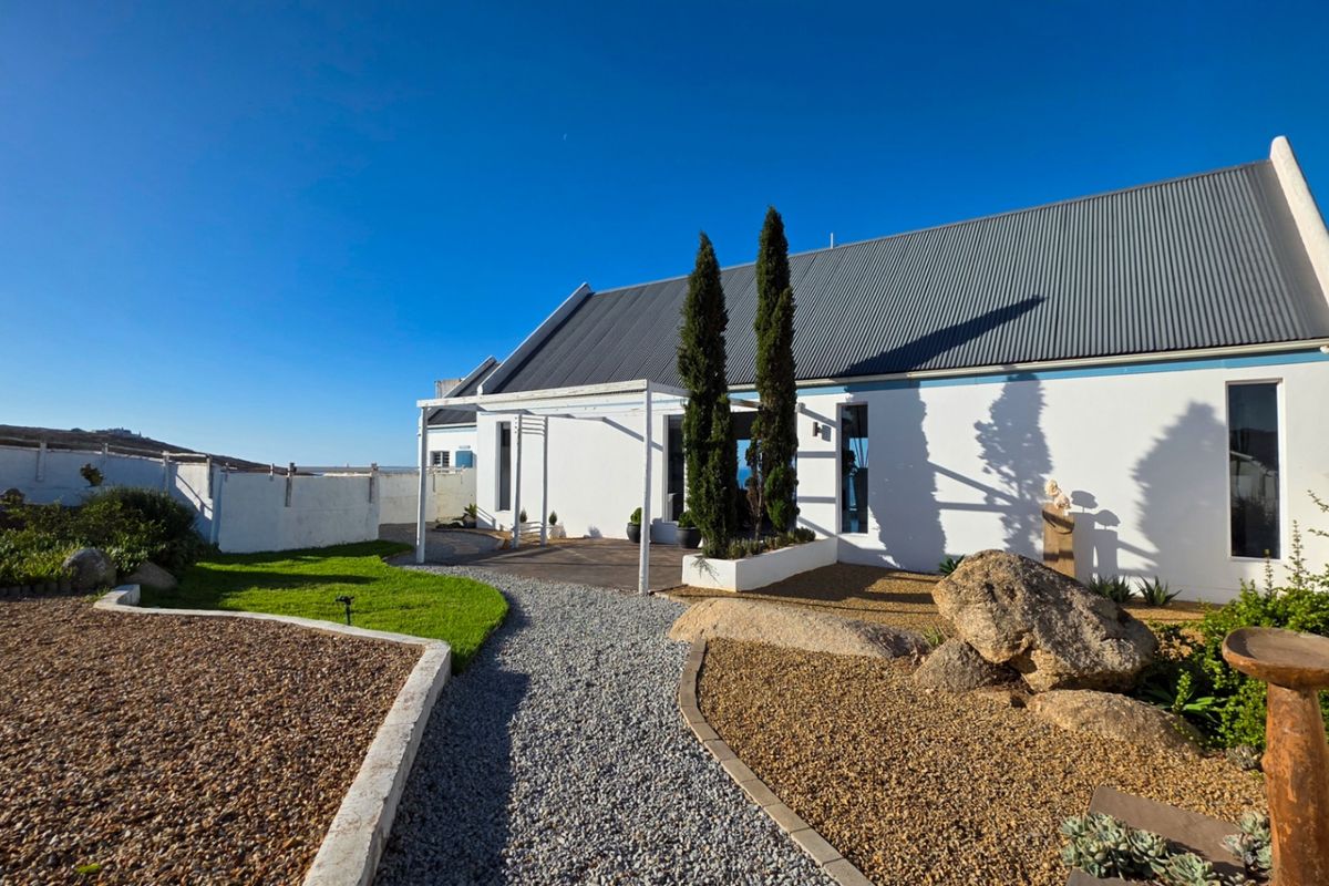 A well-maintained rear exterior view featuring a white facade, corrugated metal roof, pergola, and landscaped garden with gravel pathways.