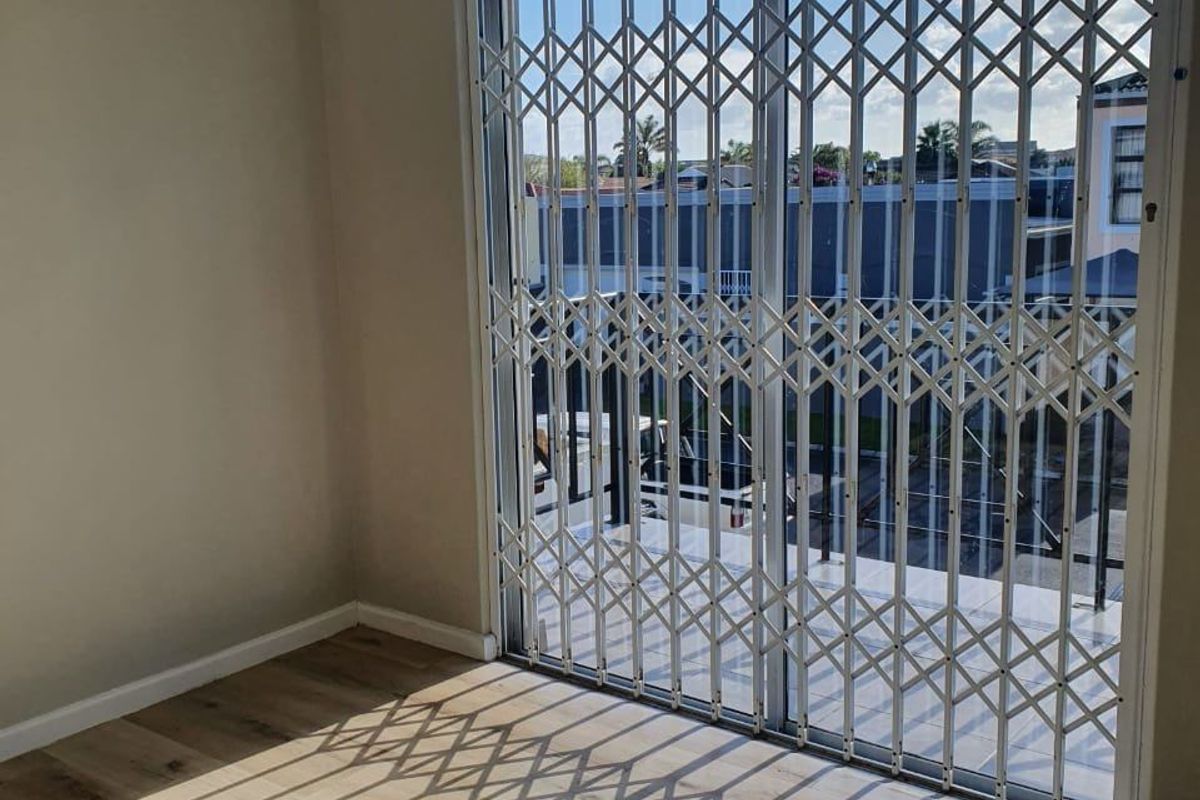 A bright bedroom featuring light wood-look flooring and a sliding glass door secured with a white metal security gate, leading to an outdoor balcony.