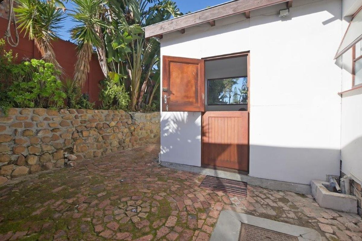 A rear exterior view featuring a stable-style timber door, brick paved courtyard, and a stone retaining wall with established garden greenery.