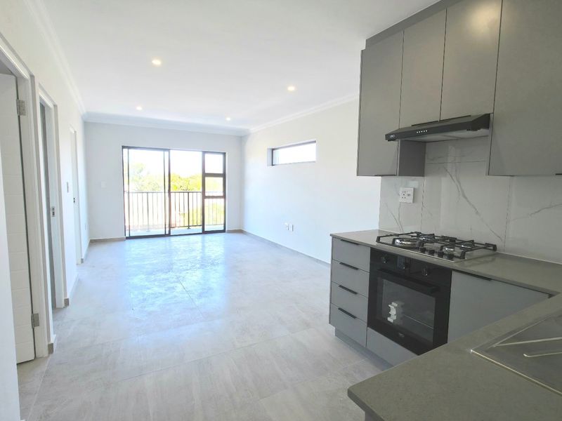 Modern open-plan living area featuring a sleek kitchen with grey cabinetry, gas hob, and stone countertops, leading to a balcony with natural light.