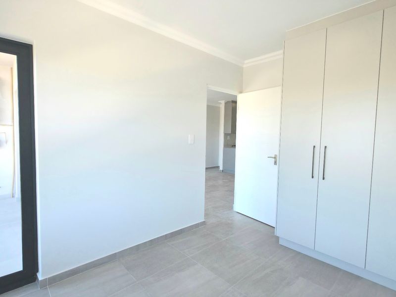 A bright, empty bedroom featuring modern built-in cupboards, neutral tiled flooring, and direct access to a balcony through a sliding door.