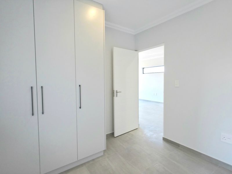 A clean, modern bedroom featuring neutral tiled flooring, built-in cupboards, and a doorway leading to an adjacent room.