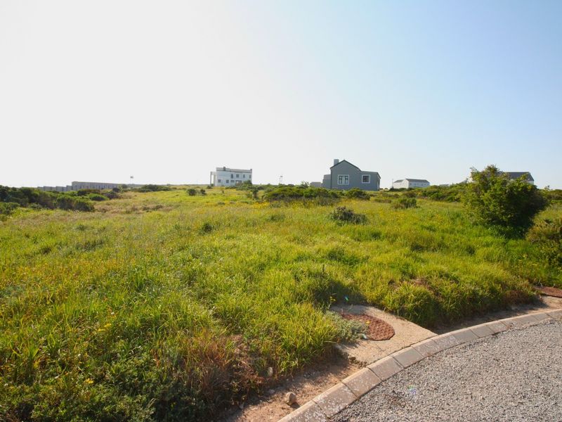 A vacant residential plot featuring natural grass coverage, accessible from a paved street with visible utility access points.