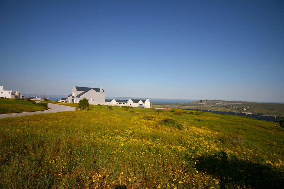 A wide-angle view of a vacant residential plot featuring natural vegetation, a paved access road, and distant coastal scenery.