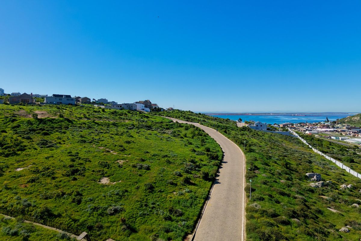 Aerial view of a vacant residential plot featuring a paved road, natural coastal vegetation, and scenic ocean views in the distance.