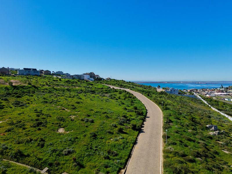 Aerial view of a vacant residential plot featuring a paved road, natural coastal vegetation, and scenic ocean views in the distance.