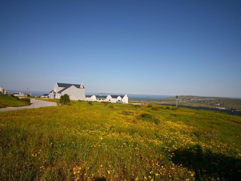 A wide-angle view of a vacant residential plot featuring natural vegetation, a paved access road, and distant coastal scenery.