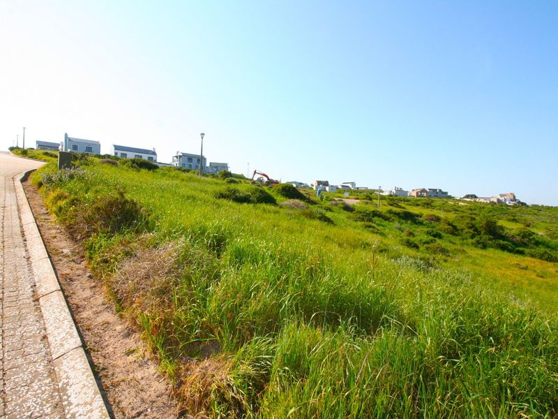 A view of the vacant residential land plot featuring natural grass coverage, adjacent to a paved road and concrete curbing in a coastal neighborhood.