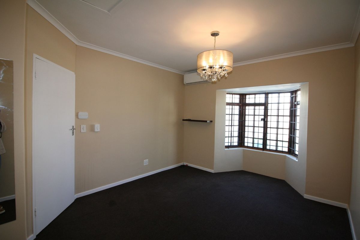 A spacious bedroom featuring a bay window with security bars, carpeted flooring, and a wall-mounted air conditioning unit.