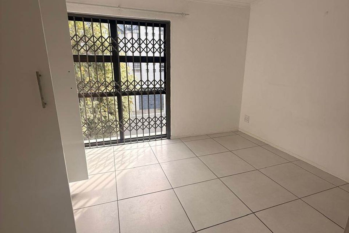 A bright, empty bedroom featuring neutral tiled flooring, a built-in wardrobe, and a window secured with a metal security gate.