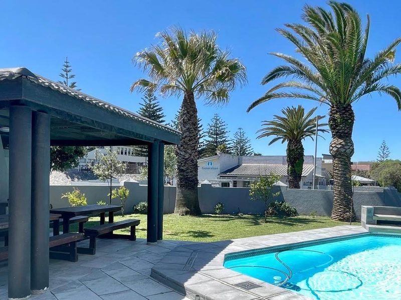 Outdoor entertainment area featuring a swimming pool, paved surround, and a covered patio with a built-in braai, set within a landscaped garden.