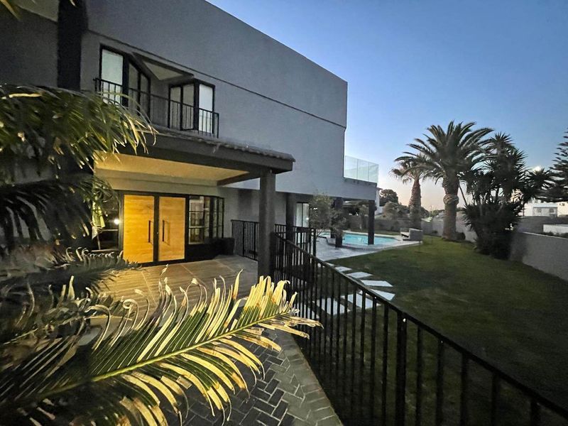 Rear view of a modern two-story house featuring a covered patio, balcony with glass balustrade, swimming pool, and a well-maintained garden.