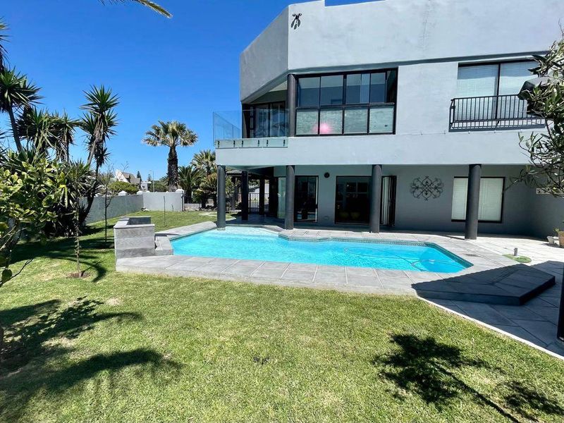 A well-maintained rear exterior featuring a swimming pool with tiled surround, glass balustrade, and a covered patio area set within a landscaped garden.