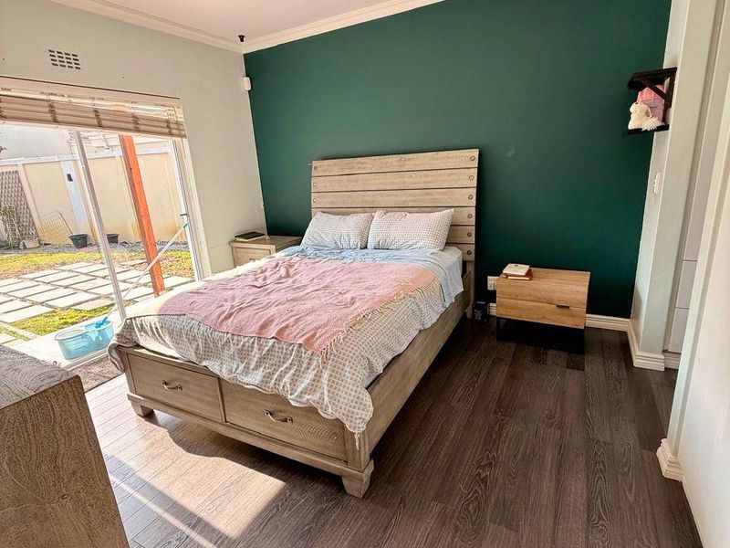 A well-lit bedroom featuring dark laminate flooring, a bold green accent wall, and a sliding glass door providing direct access to the outdoor patio area.