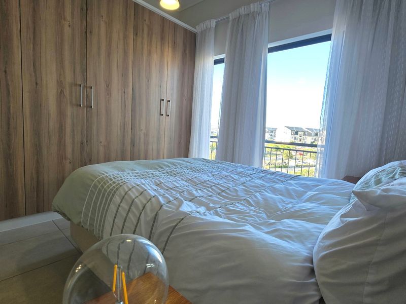 A well-lit bedroom featuring extensive built-in wooden cupboards, tiled flooring, and large windows providing natural light and access to a balcony.