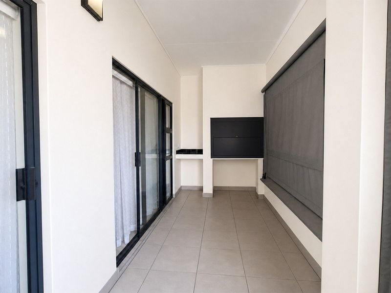A clean, modern covered balcony featuring tiled flooring, built-in braai, and integrated outdoor roller blinds, providing a versatile space for outdoor entertaining.