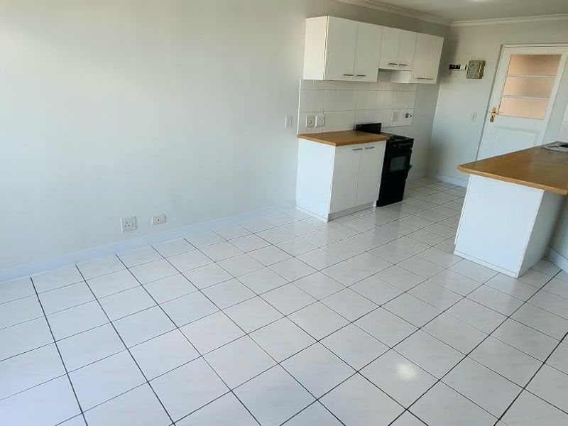 An open-plan kitchen and living area featuring white tiled flooring, built-in cabinetry with laminate countertops, and a sliding door leading to the exterior.