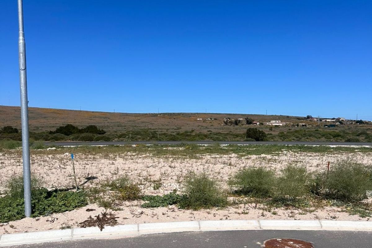 A vacant residential plot featuring a paved road frontage, concrete curbing, and utility access, offering an open landscape with distant hillside views.