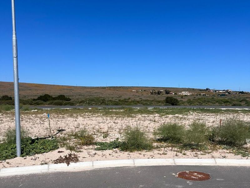 A vacant residential plot featuring a paved road frontage, concrete curbing, and utility access, offering an open landscape with distant hillside views.