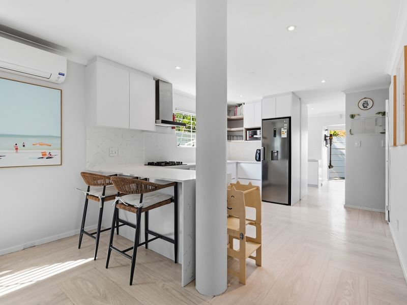 Modern open-plan kitchen featuring sleek white cabinetry, stone countertops, integrated appliances, and a central structural column, finished with light wood-look flooring.