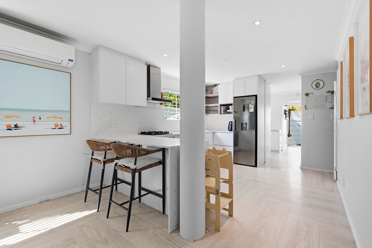 Modern open-plan kitchen featuring sleek white cabinetry, stone countertops, integrated appliances, and a central structural column, finished with light wood-look flooring.