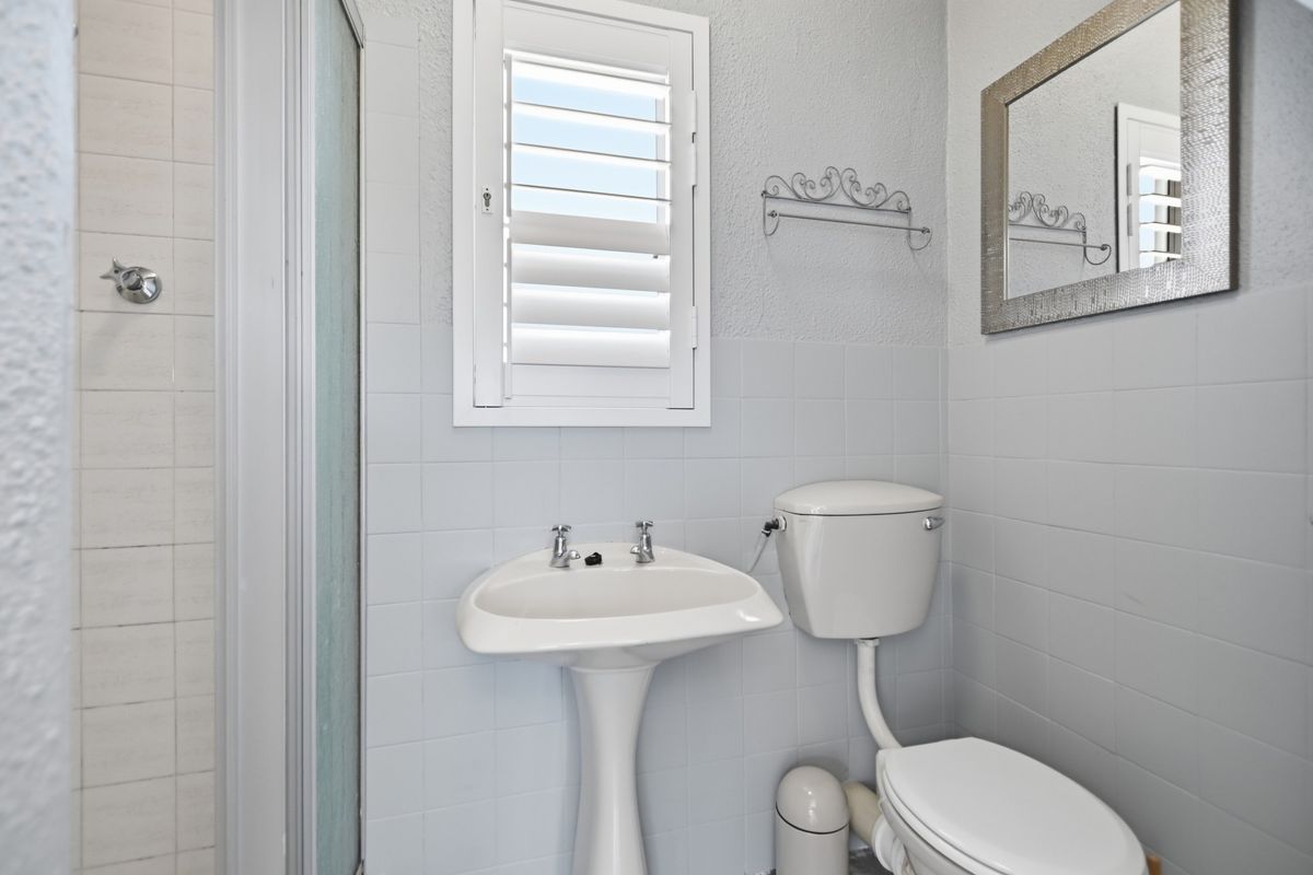 A functional bathroom featuring a pedestal basin, toilet, shower enclosure, and tiled walls with a shutter window for natural light.