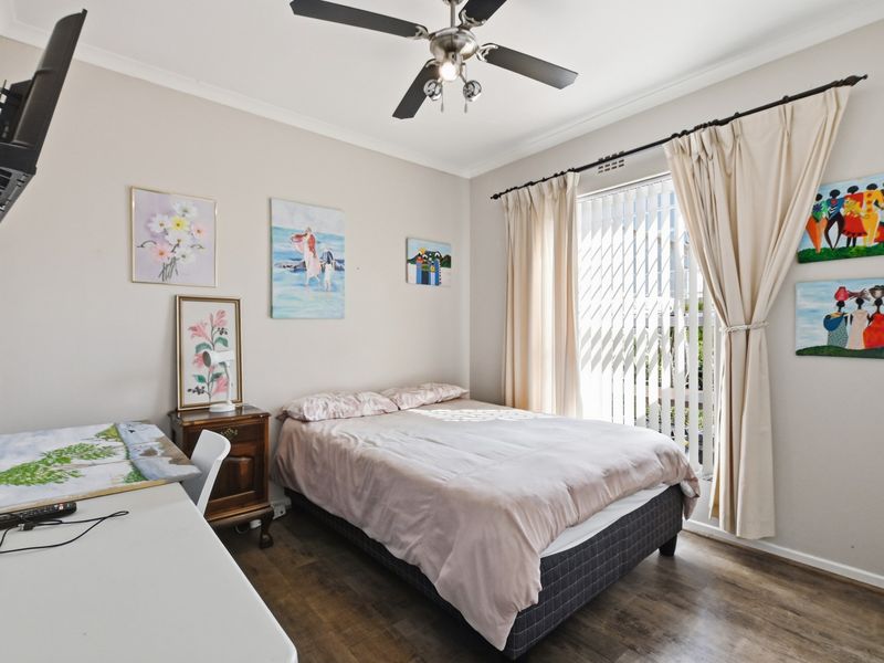 A well-lit bedroom featuring laminate flooring, a ceiling fan, and large windows with vertical blinds and curtains.