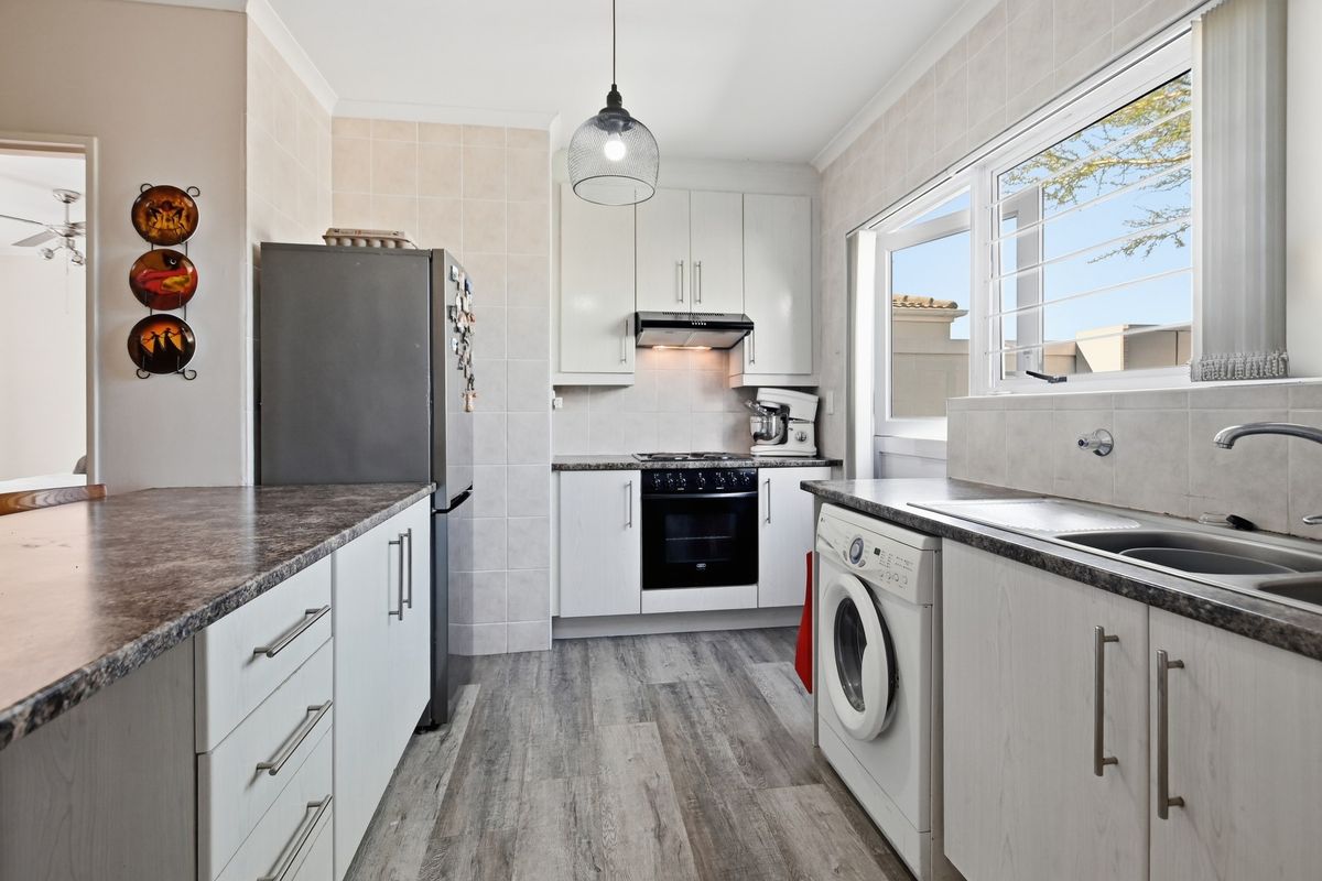 A functional kitchen featuring white cabinetry, laminate countertops, built-in oven and hob, and integrated laundry plumbing, with tiled splashbacks and wood-look flooring.