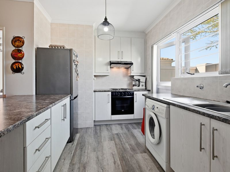 A functional kitchen featuring white cabinetry, laminate countertops, built-in oven and hob, and integrated laundry plumbing, with tiled splashbacks and wood-look flooring.