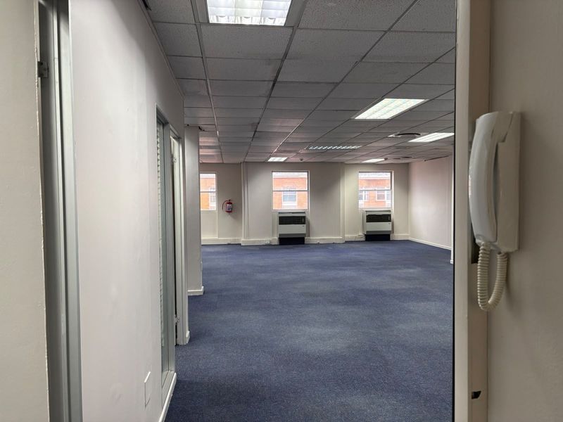 An open-plan commercial office space featuring blue carpet flooring, suspended ceilings with recessed lighting, and wall-mounted air conditioning units.