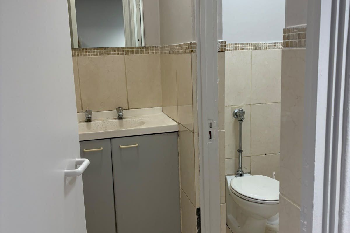 A functional commercial bathroom featuring a vanity with basin, wall mirror, and a separate toilet cubicle with tiled finishes throughout.