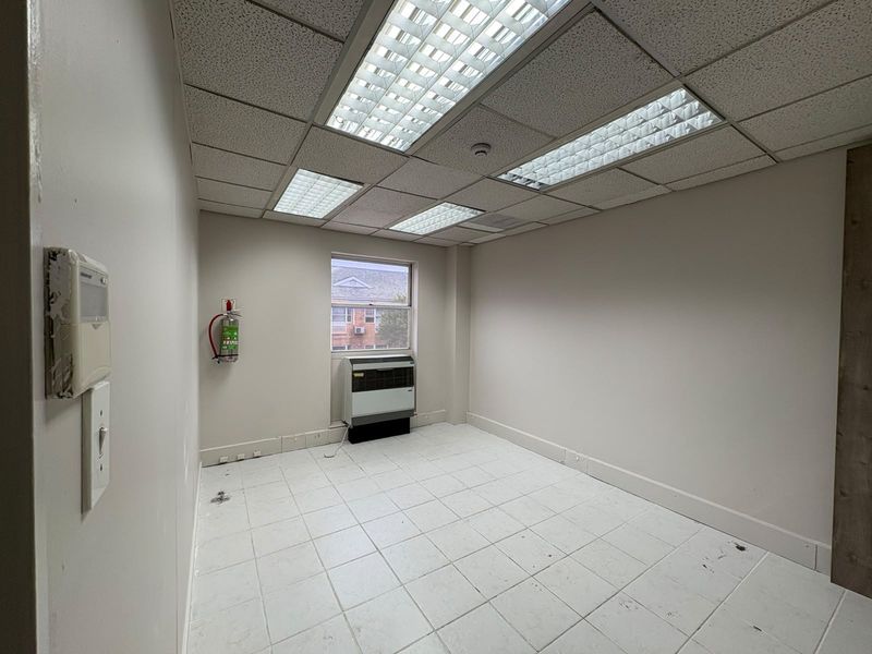 A vacant commercial office space featuring tiled flooring, a suspended ceiling with recessed lighting, and a built-in PTAC air conditioning unit.