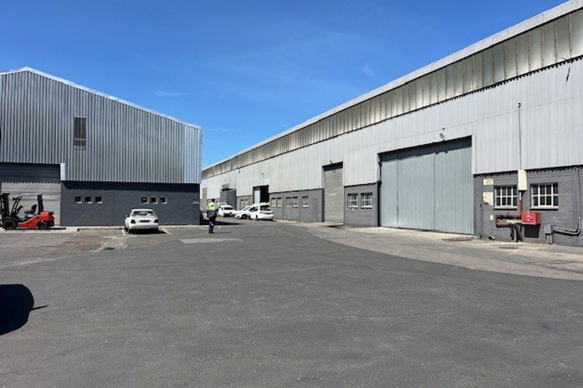 Exterior view of a commercial industrial complex featuring large metal-clad warehouse structures, roller shutter doors, and a paved asphalt yard area.