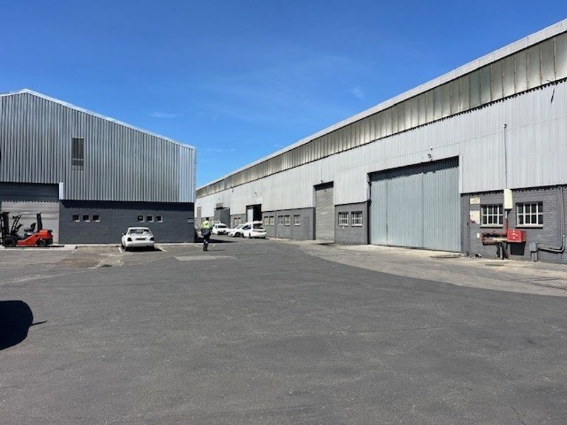 Exterior view of a commercial industrial complex featuring large metal-clad warehouse structures, roller shutter doors, and a paved asphalt yard area.