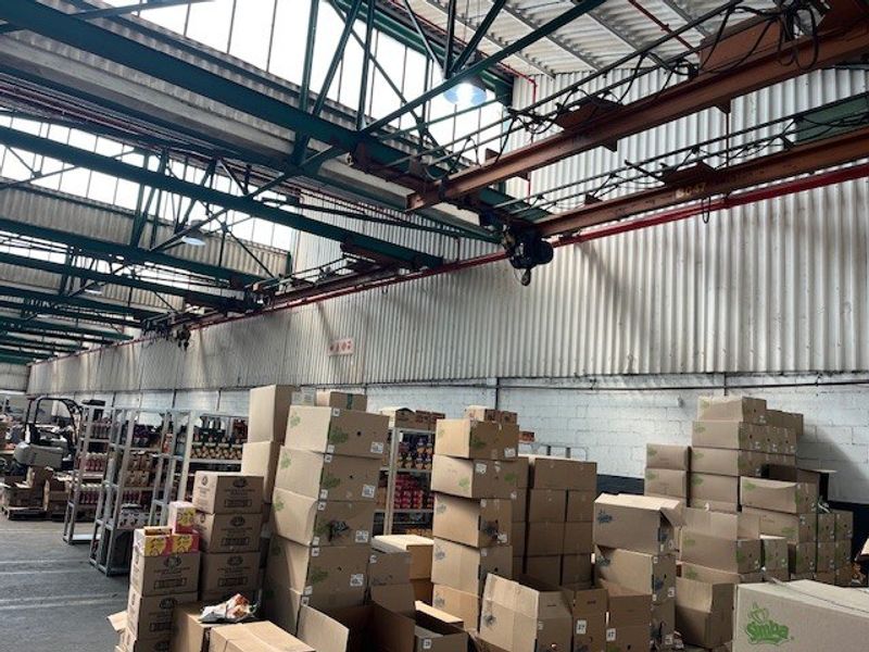 Large industrial warehouse space featuring high ceilings with exposed steel trusses, overhead crane infrastructure, and corrugated metal wall cladding.