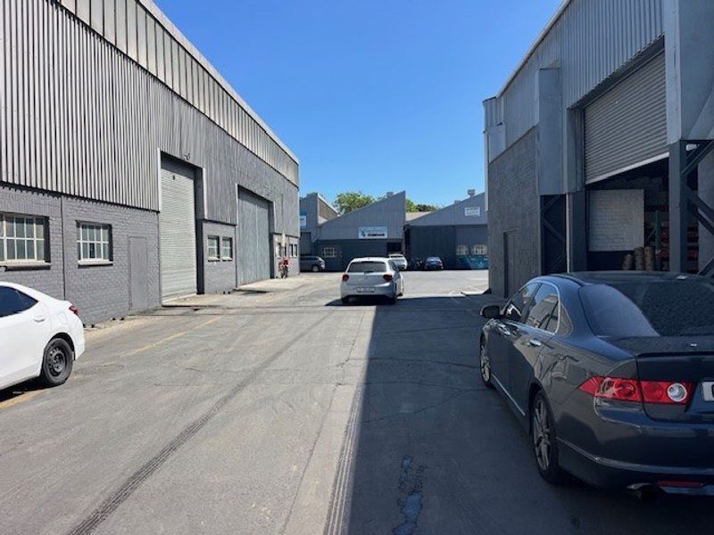 Exterior view of a commercial industrial complex featuring corrugated metal warehouse buildings, roller shutter doors, and an asphalt access driveway.