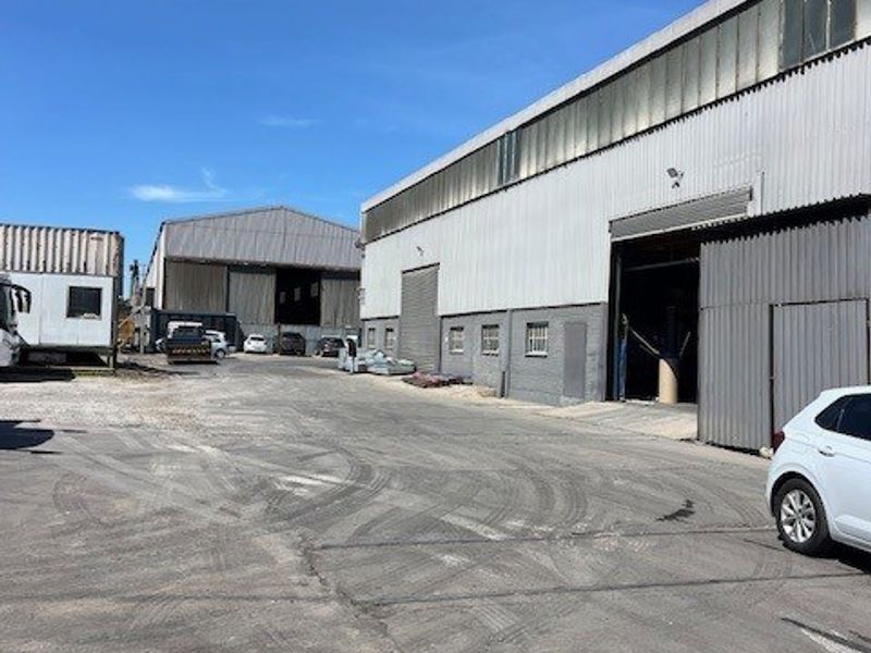 Exterior view of a commercial industrial warehouse facility featuring corrugated metal cladding, large roller shutter access doors, and a paved asphalt yard area.