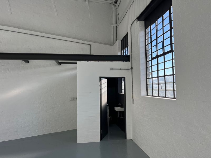 Spacious industrial-style open plan area featuring high ceilings, exposed steel beams, large steel-framed windows, and polished epoxy flooring with an integrated bathroom.