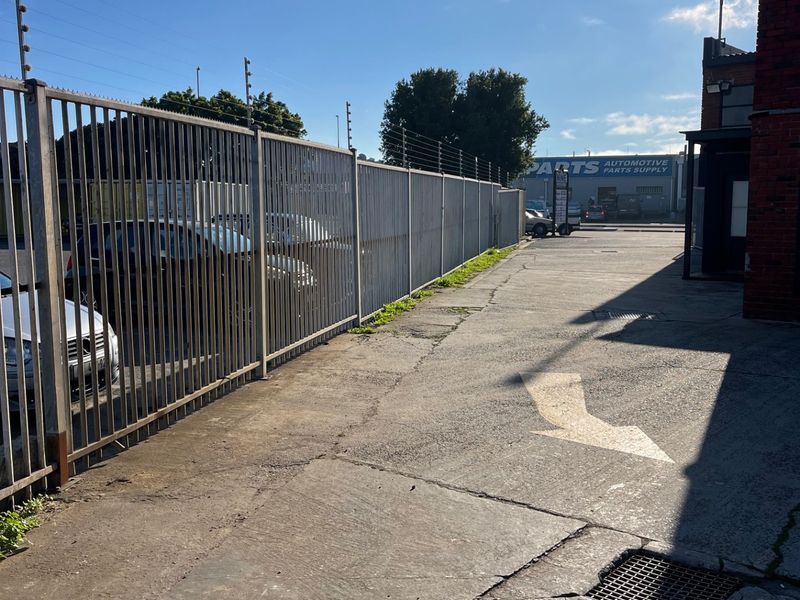 Rear commercial access area featuring concrete paving, steel palisade security fencing with electric wires, and a drainage grate alongside a brick building structure.