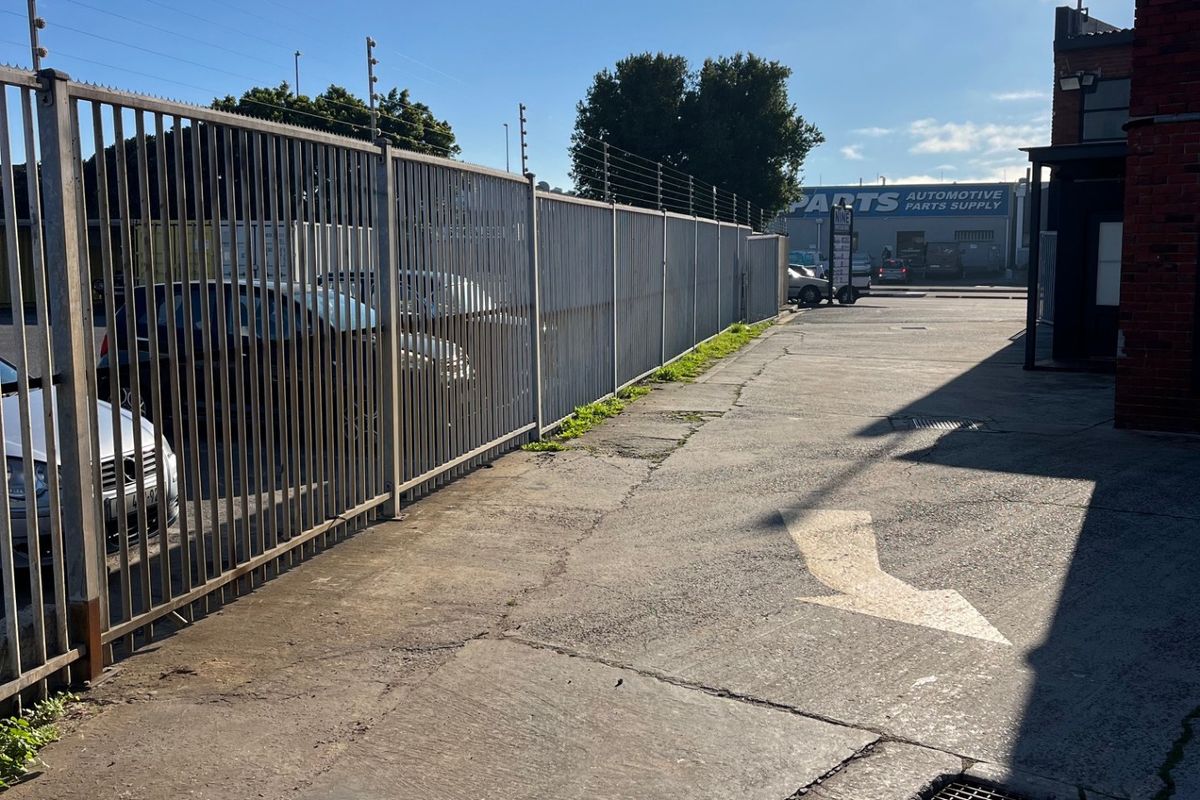 Rear commercial access area featuring concrete paving, steel palisade security fencing with electric wires, and a drainage grate alongside a brick building structure.
