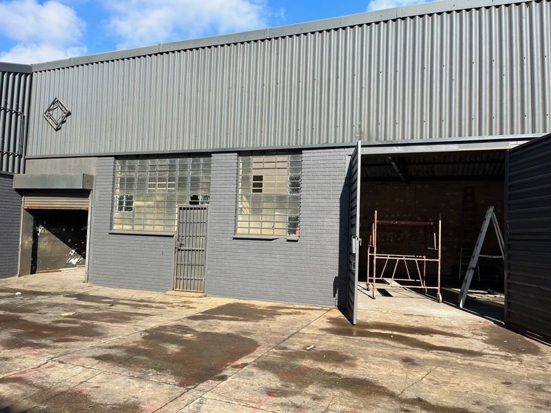 Commercial industrial facade featuring corrugated metal cladding, brickwork, security gates, and roller shutter access points with a concrete apron.